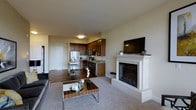 This single-floor residence offers a thoughtfully designed open-concept layout that seamlessly connects the living and kitchen areas, creating a functional and inviting atmosphere. The living area is anchored by a striking white-mantled fireplace, which serves as the room's centerpiece and adds a warm, elegant touch. A flat-screen television is mounted above the mantle, maximizing both style and practicality. The space features a combination of plush carpeting in the living area and rich hardwood-style flooring in the kitchen and entryway zones, providing a clear and visually appealing delineation between spaces. The kitchen is well-equipped with dark wood cabinetry, stainless steel appliances including a refrigerator, microwave, and dishwasher, and ample counter space that makes it ideal for everyday cooking. A utility shelf near the kitchen entrance adds convenient additional storage. Natural light fills the space through the open layout, complemented by recessed and flush-mount ceiling lighting. The entryway, visible at the far end of the space, features a clean white door and smooth transition into the main living areas. Overall, the residence presents a balanced blend of modern finishes and comfortable design.