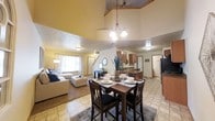 This two-floor home is designed around an open-concept main living area that seamlessly connects the dining room, living room, and kitchen into one cohesive space. The most striking architectural feature is the dramatic vaulted ceiling that rises above the living and dining areas, creating an impressive sense of height and volume. A stylish chandelier hangs above the dining area, which centers around a dark wood table with seating for four, complemented by a patterned area rug beneath. The living room is carpeted and bathed in natural light from a large window dressed with decorative curtains, furnishing a comfortable and welcoming atmosphere. The kitchen is positioned just beyond the dining area and features warm wood cabinetry, tile flooring, and ample counter space, with direct sightlines into the main living areas. Tile flooring flows through the dining and kitchen spaces, providing a clean, durable surface that contrasts nicely with the carpeted living room. The layout of the main floor encourages an open flow between everyday living spaces, while a staircase leads to the second floor, expanding the overall footprint of the home. The warm, neutral color palette throughout contributes to a welcoming and lived-in ambiance.