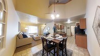 This two-floor home is designed around an open-concept main living area that seamlessly connects the dining room, living room, and kitchen into one cohesive space. The most striking architectural feature is the dramatic vaulted ceiling that rises above the living and dining areas, creating an impressive sense of height and volume. A stylish chandelier hangs above the dining area, which centers around a dark wood table with seating for four, complemented by a patterned area rug beneath. The living room is carpeted and bathed in natural light from a large window dressed with decorative curtains, furnishing a comfortable and welcoming atmosphere. The kitchen is positioned just beyond the dining area and features warm wood cabinetry, tile flooring, and ample counter space, with direct sightlines into the main living areas. Tile flooring flows through the dining and kitchen spaces, providing a clean, durable surface that contrasts nicely with the carpeted living room. The layout of the main floor encourages an open flow between everyday living spaces, while a staircase leads to the second floor, expanding the overall footprint of the home. The warm, neutral color palette throughout contributes to a welcoming and lived-in ambiance.