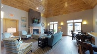 This single-floor space makes an immediate impression with its dramatic, vaulted ceiling finished in warm, natural wood planking that stretches across the entire room, creating a striking architectural focal point. The open-concept layout seamlessly connects the living and dining areas, fostering a sense of spaciousness and flow throughout.

The living area is anchored by a classic fireplace set into a bold, dark accent wall, above which a wall-mounted television is positioned for comfortable viewing. Seating is arranged thoughtfully around a tufted ottoman coffee table, with teal upholstered sofas and patterned accent chairs providing both style and comfort. A decorative area rug grounds the seating arrangement on the light hardwood flooring that runs throughout the space.

Adjacent to the living area, a dining space features a dark wood table and chairs, bathed in natural light from large windows and glass doors that offer views of and direct access to an outdoor patio area. Wall sconces and recessed lighting complement the natural light, creating a warm, inviting ambiance throughout. The overall design balances rustic warmth with modern, transitional style.