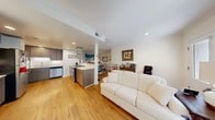 Located at 2217 Rogene Dr in Baltimore, MD, this efficiently designed single-floor residence offers a thoughtfully organized layout that maximizes every square foot. The open-concept living, dining, and kitchen area forms the heart of the home, unified by warm hardwood flooring throughout. The kitchen is a standout feature, boasting contemporary gray cabinetry, crisp white quartz countertops, stainless steel appliances including a dishwasher and gas range, and a striking stainless steel island hood. A breakfast bar with seating provides a casual dining option, while recessed lighting keeps the space bright and welcoming. The dining area is defined by crown molding, a decorative chandelier, and a framed wall mirror that adds depth to the room. The living room benefits from natural light streaming through windows that frame lush greenery outside, complemented by recessed and pendant lighting. A hallway connects the main living spaces to the full bathroom, which features tile flooring and a shower-tub combination. The dedicated laundry room includes a full-sized washer and dryer with utility shelving, adding practical convenience. The overall ambiance is clean, modern, and comfortable, making this a functional and inviting single-floor residence.