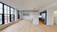 This single-floor unit showcases a thoughtfully designed open-concept layout that blends industrial and contemporary elements throughout. Light-toned hardwood flooring extends seamlessly across the living and kitchen areas, contributing to a bright, cohesive aesthetic. Exposed cylindrical ductwork runs overhead, adding an industrial character that complements the otherwise clean, white interior.

The kitchen is a standout feature, anchored by a large island with a white quartz countertop, an integrated sink, and a dishwasher. Navy-blue cabinetry contrasts sharply against the white surfaces, while stainless steel appliances—including a gas range, microwave, and French door refrigerator—complete the functional setup. Pendant lights hang above the island, providing focused task lighting.

The living area benefits from generous natural light streaming through oversized black-framed multi-pane windows, which frame views of the surrounding urban streetscape. Floating wood shelves add a subtle decorative touch to the main living space. The bedroom mirrors this aesthetic, featuring similar black-framed windows, a ceiling fan, and ample floor space.

The bathroom is finished with white hexagonal mosaic tile on the walls surrounding the soaking tub and shower, contrasted by large-format gray floor tiles. In-unit laundry with a stacked washer and dryer adds everyday convenience.