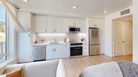 Located at 414 Front St in downtown Santa Cruz, this thoughtfully designed studio offers a efficient, modern layout across a single floor. The open-plan kitchen and living area serves as the heart of the space, featuring sleek white cabinetry extending to the ceiling, a textured tile backsplash, stainless steel appliances including a dishwasher, range, over-the-range microwave, and refrigerator, and light quartz countertops. Light wood-tone flooring flows throughout, enhancing the clean, contemporary aesthetic. The living area is bathed in natural light through large floor-to-ceiling windows that frame views of the surrounding downtown streetscape, with sheer curtains adding a soft, airy touch. The bedroom area is accented by warm neutral tones and provides ample closet storage. A dedicated workspace features a marble-topped vanity desk with a bold black-framed mirror, blending style with function. The full bathroom is finished in crisp white tile, complete with a soaking tub and shower combination, a white vanity with black hardware, and warm wood-tone flooring. An in-unit laundry area and two closets provide practical everyday convenience. The overall ambiance is bright, clean, and contemporary throughout.