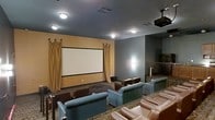 This private screening room offers an immersive cinematic experience designed for comfort and entertainment. The focal point of the space is a large projection screen mounted on a warm amber-toned accent wall, framed by elegant floor-to-ceiling draping curtains that enhance the theatrical ambiance. A ceiling-mounted projector ensures a crisp, high-quality viewing experience from every seat in the room.

The seating is arranged in a tiered layout to provide unobstructed sightlines throughout. Guests can choose from a combination of plush blue-gray armchairs positioned toward the front and rich brown leather recliners lining the rear rows, accommodating a generous number of viewers in style. The patterned carpeted flooring adds a classic, upscale cinema feel underfoot while also contributing to the room's acoustics.

Ambient lighting is thoughtfully integrated into the ceiling, providing subtle illumination that complements the viewing environment without creating glare. The gray side walls keep the focus directed toward the screen, while aisle lighting and accent fixtures add depth to the overall atmosphere. A staircase visible along one side indicates tiered access between seating levels. This well-appointed screening room is an ideal venue for movie nights, presentations, or group entertainment.