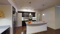 This single-floor space is anchored by a beautifully designed open-concept kitchen that serves as the heart of the home. Rich dark espresso cabinetry lines the walls, offering ample storage while creating a striking contrast against the light-toned walls and gleaming granite countertops. The kitchen is equipped with a full suite of stainless steel appliances, including a refrigerator, range with gas cooktop, and built-in microwave, making it as functional as it is stylish. A curved peninsula breakfast bar extends the counter space and provides a seamless transition between the kitchen and the adjacent living area, ideal for casual dining or entertaining. Overhead, a combination of track lighting and elegant pendant lights illuminate the workspace with a warm, inviting glow. The floors throughout are finished in rich hardwood, adding warmth and a sense of continuity to the open layout. Subtle tile backsplash detailing behind the range adds texture and visual interest to the cooking area. The overall ambiance is modern and sophisticated, with thoughtful design choices that balance aesthetics and practicality throughout the space.