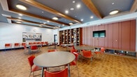 Located at 3223 Timber Mill Pkwy in Castle Rock, Colorado, this single-floor community room is a well-appointed gathering space designed for social interaction and group activities. The room accommodates multiple round tables paired with vibrant red modern chairs, creating a welcoming and functional layout throughout the open floor plan.

Architectural interest is provided by the striking ceiling design, which features exposed wooden beams set against a deep slate-blue backdrop, complemented by recessed lighting and flush drum fixtures that ensure the space is well-lit throughout. The warm-toned tile flooring adds texture and durability underfoot.

A prominent built-in shelving unit spans a significant portion of one wall, displaying decorative ceramics, vases, and sculptural objects that contribute an artistic character to the room. Adjacent to the shelving, a richly toned wood-paneled accent wall serves as the backdrop for a wall-mounted flat-screen television, with additional lower shelving providing storage for books and materials.

Large windows along one wall allow natural light to fill the space, offering views of the surrounding exterior. A set of double glass doors connects the room to adjacent areas, enhancing the sense of connectivity within the broader facility. The overall ambiance is modern yet inviting, suitable for community events, meetings, or casual gatherings.