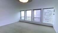 This single-floor space offers a clean, minimalist aesthetic defined by its crisp white walls, soft carpeted flooring, and an abundance of natural light. The most commanding feature is the expansive bank of large windows that lines one wall, framing breathtaking elevated views of the waterfront, a scenic green esplanade, and the surrounding urban skyline. The windows flood the interior with daylight, creating an open, airy atmosphere throughout the space. A flush ceiling light fixture provides supplemental illumination, maintaining the room's understated and modern character. The neutral color palette — from the off-white walls to the light beige carpet — gives the space a versatile, move-in-ready feel that can accommodate a variety of uses and furnishing styles. The elevated vantage point suggests a high-floor position within a larger building, making the panoramic outdoor views a defining and exceptional characteristic of this space. Overall, the interior exudes a calm, professional ambiance that balances urban connectivity with a sense of tranquility, making it well-suited for residential or professional use.