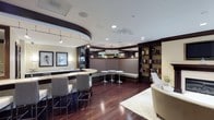 This elegantly designed single-floor space combines modern sophistication with functional versatility, making it well-suited for socializing, networking, or quiet reading. The centerpiece is a curved bar counter with granite or stone-top surfaces and stylish bar seating, anchored by a distinctive circular ceiling detail with recessed and pendant lighting that adds architectural drama overhead. Rich, dark hardwood flooring flows throughout, grounding the space with warmth and a polished aesthetic. A built-in banquette seating area with high backs lines one side of the room, accompanied by sleek round pedestal tables — ideal for casual conversation or small group gatherings. On the opposite side, floor-to-ceiling dark wood built-in bookshelves are stocked with books and decorative accents, lending the space a refined, library-like quality. A cozy lounge area with a contemporary armchair and a soft area rug provides an inviting retreat for more relaxed seating. The lighting throughout is thoughtfully layered, combining recessed ceiling lights, pendant fixtures, and wall sconces to create a warm, welcoming ambiance. The overall design palette of cream, taupe, and deep espresso tones conveys a sense of upscale comfort and understated luxury.