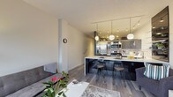 This contemporary single-floor residence at 1224 West Riverside Avenue in Spokane, Washington, offers a thoughtfully designed open-concept layout with modern finishes throughout. The kitchen is a standout feature, showcasing high-gloss dark cabinetry, stainless steel appliances including a French door refrigerator, a mosaic tile backsplash, and a generous marble-top island with bar seating — all illuminated by striking globe pendant lights and track lighting overhead. Glass shelving adds a refined, open display element to the kitchen's design. The living area flows naturally from the kitchen and is anchored by a fireplace set against a bold teal accent wall. Floor-to-ceiling corner windows flood the space with natural light and frame lush treetop views, while a sliding glass door provides access to an outdoor balcony. Light wood-style flooring runs consistently throughout, lending warmth and continuity to the space. A hallway with sleek glass-panel sliding doors leads to the bedroom, which is comfortably proportioned and well-lit. The bathroom is finished with large-format white wall tiles, a frameless glass sliding shower enclosure with a mosaic tile floor, and a clean white vanity with modern fixtures. Track lighting and thoughtful design details are present throughout, reflecting a cohesive, urban aesthetic.