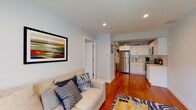 Located at 5959 Broadway in the Riverdale neighborhood of New York City, this single-floor residence offers a thoughtfully designed layout with a cohesive, modern aesthetic throughout. Rich hardwood flooring flows continuously across the living spaces, lending warmth and visual continuity to the entire unit.

The open-plan living area is bright and inviting, painted in soft blue-gray tones that create a calm atmosphere. A large wall-mounted flat-screen TV anchors the room, complemented by a comfortable sofa, a zebra-print area rug, and a wooden coffee table. Natural light enters through a window dressed with patterned curtains, adding depth to the space.

The kitchen is efficiently designed with white shaker-style cabinetry, light subway tile backsplash, white countertops, and a full suite of stainless steel appliances including a refrigerator, electric range, and over-the-range microwave. Hardwood flooring continues seamlessly into this area.

The primary bedroom features a tufted upholstered bed frame, a natural wood-framed floor mirror, and a window that admits natural light. A secondary bedroom includes a wall-mounted TV and a window with blinds and curtains.

The bathroom is fully renovated with floor-to-ceiling white subway tile, a tub-shower combo with a sliding glass door, a white vanity with mirror cabinet, and marble-look tile flooring — clean and polished in design. The entryway is anchored by a solid wood door and provides direct access to the bathroom.