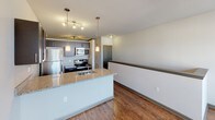 This single-floor space is designed with a modern, open-concept layout that seamlessly connects the kitchen and living areas. The kitchen serves as a central hub, showcasing granite countertops with subtle speckled detailing and a large peninsula that doubles as a breakfast bar. Dark espresso-toned cabinetry provides strong visual contrast against the light countertops, while stainless steel appliances—including a refrigerator, range, and microwave—add a sleek, contemporary touch. Pendant lighting hangs above the peninsula, complemented by a track lighting system that illuminates the cooking area effectively.

Hardwood-style flooring runs throughout the main living spaces, adding warmth and cohesion to the open floor plan. The living area benefits from a generous bank of windows that allow ample natural light to fill the room, with exterior views of the surrounding neighborhood. A ceiling fan with a built-in light fixture provides both comfort and additional illumination.

The bathroom features a full tub and shower combination with a curved chrome rod and clean white surround, finished with neutral-toned walls for a simple, functional aesthetic. Overall, the space presents a clean, contemporary atmosphere with practical finishes suited for everyday living.