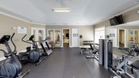 This fitness center is a comprehensive, single-floor facility designed to support a full range of workout routines. The space is divided into distinct functional zones, each thoughtfully appointed for a specific type of exercise.

The main gym area features durable rubber flooring and is lined with large windows fitted with white blinds, allowing ample natural light to fill the space. Wall-mounted mirrors expand the visual footprint of the room and allow users to monitor their form. Overhead, crown molding lends a refined architectural detail throughout. Multiple flat-screen televisions are mounted near the ceiling, providing entertainment during workouts.

The strength training section is equipped with a variety of weight machines, a dumbbell rack with a full range of free weights, and adjustable benches, offering options for both isolation and compound exercises. The cardio area houses upright stationary bikes, a recumbent bike, and treadmills, all positioned in front of generous windows that provide natural light and an airy atmosphere.

A separate studio room serves as a dedicated space for flexibility and mind-body training. It features the same rubber flooring, mirrored walls, exercise balls in multiple sizes, yoga mats, and balance equipment, making it ideal for yoga, Pilates, or stretching sessions. The overall ambiance is clean, bright, and well-maintained.