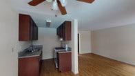 This single-floor residence offers a practical and comfortable layout with a seamlessly connected kitchen and living area. The kitchen is outfitted with rich, dark wood cabinetry that extends to the upper walls, providing generous storage space. Granite-style countertops line the workspace, complemented by a stainless steel sink and a freestanding gas range, making the kitchen both functional and visually appealing. A structural column subtly defines the boundary between the kitchen and the adjoining living area, maintaining an open and airy feel throughout. Warm-toned hardwood-style flooring runs continuously across both spaces, adding cohesion and a sense of warmth to the interior. A ceiling fan with wooden blades is centrally positioned, enhancing air circulation and contributing to the room's relaxed aesthetic. Neutral-toned walls provide a clean, versatile backdrop throughout. A hallway visible from the main living space suggests additional rooms beyond, completing the functional flow of this well-appointed single-level home.