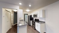 This well-appointed single-floor residence offers a thoughtfully designed layout that balances functionality and modern style. The kitchen serves as a focal point of the space, showcasing crisp white shaker-style cabinetry that extends to the upper walls, providing ample storage. Dark countertops create a striking contrast against the white cabinets, while a subway tile backsplash adds a clean, contemporary touch. Stainless steel appliances — including a refrigerator, range, and over-the-range microwave — lend a polished, professional feel to the cooking area. A kitchen peninsula with an integrated sink offers additional prep space and casual seating potential, making it an ideal hub for both cooking and entertaining. Recessed lighting illuminates the kitchen evenly, enhancing the bright and open atmosphere. Rich, hardwood-style flooring runs throughout, adding warmth and visual continuity to the space. A walk-in closet is visible beyond the kitchen, offering generous storage. Accent walls in deep charcoal grey provide depth and contrast against the predominantly neutral palette, contributing to the space's modern and inviting ambiance.