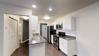 This well-appointed single-floor residence offers a thoughtfully designed layout that balances functionality and modern style. The kitchen serves as a focal point of the space, showcasing crisp white shaker-style cabinetry that extends to the upper walls, providing ample storage. Dark countertops create a striking contrast against the white cabinets, while a subway tile backsplash adds a clean, contemporary touch. Stainless steel appliances — including a refrigerator, range, and over-the-range microwave — lend a polished, professional feel to the cooking area. A kitchen peninsula with an integrated sink offers additional prep space and casual seating potential, making it an ideal hub for both cooking and entertaining. Recessed lighting illuminates the kitchen evenly, enhancing the bright and open atmosphere. Rich, hardwood-style flooring runs throughout, adding warmth and visual continuity to the space. A walk-in closet is visible beyond the kitchen, offering generous storage. Accent walls in deep charcoal grey provide depth and contrast against the predominantly neutral palette, contributing to the space's modern and inviting ambiance.