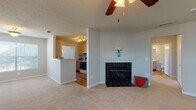 This single-floor home offers a well-organized layout with distinct living, kitchen, and bedroom areas. The spacious living room is anchored by a black tile-surround fireplace with a white mantel, complemented by soft neutral carpet and a ceiling fan overhead. A textured ceiling adds a classic touch, while a large window invites natural light and provides views of an outdoor balcony/patio area accessible through a glass-panel door.

The kitchen follows a galley-style configuration, featuring light wood cabinetry, white countertops, hardwood flooring, and white appliances including a range and dishwasher. A pass-through breakfast bar connects the kitchen to the living area, and a doorway leads to a dedicated laundry space illuminated by a distinctive octagonal window.

The bedroom is generously proportioned with plush carpet, a ceiling fan, and direct access to a full bathroom. A spacious walk-in closet with wire shelving provides ample storage. Two bathrooms are present throughout the home: one full bath with a tub/shower combination and hardwood floors, and a second bathroom featuring a single vanity, large mirror, and warm overhead lighting. Both bathrooms include light wood cabinetry and neutral countertops, maintaining a cohesive aesthetic throughout the home.