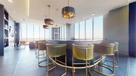 This single-floor high-rise amenity space delivers an elevated, hotel-inspired atmosphere defined by contemporary design and expansive city views. Floor-to-ceiling glass curtain walls dominate multiple areas of the space, flooding the interiors with abundant natural light and framing dramatic panoramic skyline vistas from nearly every vantage point.

At the heart of the space is a curved wet bar finished in deep tones and surrounded by plush velvet bar stools with brass-finished bases, set atop a navy hexagonal tile floor. Overhead, statement pendant lights with matte black drum shades and brass accents provide warm, layered illumination. Adjacent to the bar, a generously proportioned lounge area features a channel-tufted sectional sofa, a wall-mounted media display, and a built-in shelving unit backed by a textured geometric accent wall.

A glass-enclosed conference or private dining room offers a more intimate setting, while a sunny seating nook along the window wall features chartreuse ottomans and a bistro table. An outdoor terrace, accessible through sliding glass doors, includes high-top tables with woven seating and overhead heaters. Wide-plank hardwood flooring flows throughout the space, unifying each distinct area into one cohesive, sophisticated environment.