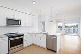 This single-floor residence is designed with a clean, modern aesthetic that emphasizes bright, open living. The entry leads into an open-concept layout where the kitchen flows seamlessly into the living area. The kitchen is well-appointed with white shaker-style cabinetry extending to the ceiling, a subway tile backsplash, light stone countertops, and a full suite of stainless steel appliances including a refrigerator, range, microwave, and dishwasher. Pendant lighting and recessed fixtures illuminate the space, while warm-toned hardwood flooring runs throughout the main living areas. Large windows and a glass door to the balcony flood the living space with natural light and offer views of the surrounding cityscape. The bedrooms are carpeted and feature built-in shelving and generous closet space, including walk-in closets with multi-level hanging rods and shelving. The primary bathroom includes a double-sink vanity with stone countertops, a walk-in tiled shower, and large mirrors with modern chrome lighting fixtures. A second full bathroom offers a single vanity, tiled tub-shower combination, and direct access to an additional walk-in closet. Throughout the residence, white walls, consistent finishes, and thoughtful storage solutions contribute to a cohesive, functional living environment.