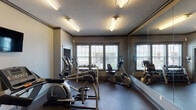 This single-floor fitness center is thoughtfully designed to support a comprehensive workout experience. The space is organized across two connected rooms, each outfitted with a dedicated selection of exercise equipment. The main area features elliptical trainers, stationary recumbent and upright bikes, and treadmills, all arranged to allow comfortable use and clear sightlines. A row of dumbbells on a tiered rack provides a free weights station, catering to strength training needs. A weight bench with an adjustable rack further supports resistance workouts. Full-length mirrors line the walls throughout, creating a sense of expanded space while allowing users to monitor their form. Multiple windows fitted with horizontal blinds invite natural light into the space and offer exterior views, enhancing the workout environment. Overhead fluorescent lighting ensures the room remains well-lit at all times. The flooring is a durable, dark rubber-style surface suited to high-traffic fitness use. Crown molding along the ceiling adds a refined architectural detail. A wall-mounted television and a decorative wall clock complete the practical amenities. The two rooms are connected by a trimmed doorway, allowing for a seamless flow between workout zones.