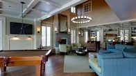 This thoughtfully designed single-floor space makes a striking impression with its soaring two-story coffered ceilings, which lend an air of grandeur and openness to the main living area. A dramatic stone fireplace serves as the room's focal point, flanked by a large ornate mirror that amplifies the sense of space and light. An elegant wrought-iron chandelier anchors the center of the room, casting a warm, inviting glow across the rich hardwood flooring below.

The open-plan layout seamlessly connects multiple functional zones. A billiards table positioned near large plantation-shuttered windows defines a dedicated recreation area, while a comfortable seating arrangement featuring green accent chairs and a blue sofa surrounds a dark circular coffee table, creating an ideal gathering space. Clerestory windows positioned high on the warm-toned walls draw in natural light from above, enhancing the airy atmosphere.

Transitioning toward the rear, built-in shelving and cabinetry provide both storage and display space, while a defined dining area with a separate chandelier adds another layer of functionality. Wall sconces throughout contribute to a layered lighting scheme that complements the space's warm, earthy color palette. The overall ambiance is one of refined comfort, blending classic architectural details with inviting, livable design.