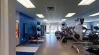 This fitness center offers a comprehensive workout environment designed to accommodate a wide range of exercise needs. The space is defined by a clean, functional layout with a drop-panel ceiling outfitted with recessed fluorescent lighting that ensures consistent, bright illumination throughout. A bold blue accent wall serves as a striking visual anchor on one side of the room, lending the space an energetic and motivating atmosphere.

Along the right side, a row of cardio machines — including treadmills and ellipticals — lines the wall, providing ample equipment for aerobic workouts. On the opposite side, a dedicated functional training zone features a tiered rack stocked with medicine balls and exercise balls in various sizes, alongside a weight bench and additional strength training equipment. Rubber and vinyl flooring in blue and grey tones defines this area, offering both safety and style underfoot.

Natural light filters in through a set of glass doors at the far end of the room, connecting the interior to an outdoor area and adding an open, airy feel to the space. A wall-mounted television is positioned for easy viewing during workouts. The overall design is practical and well-organized, making this fitness center a fully functional facility suited for both cardiovascular and strength training routines.