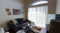 This single-floor space showcases a thoughtfully designed living area that immediately draws attention with its impressive architectural details. The living room features soaring ceilings that amplify the sense of openness, crowned by a striking arched window above a large sliding glass door fitted with vertical blinds. This combination allows abundant natural light to flood the interior while offering views of the lush outdoor greenery beyond the patio. The warm, neutral wall tones create a welcoming backdrop that complements the rich, dark leather sectional sofa and glass-top coffee table at the heart of the room. A textured area rug anchors the seating arrangement, adding depth and warmth to the space. Decorative accents, including candle holders and vibrant artwork on the walls, contribute a personalized and lived-in character. The sliding door provides direct access to an outdoor area, seamlessly blending indoor and outdoor living. Overall, the space balances comfort and style, with its architectural highlights and bright atmosphere making it a functional and aesthetically pleasing environment suited for both relaxing and entertaining.