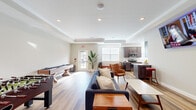 Located at 100 Grenache Blanc Dr in Saint Peters, Missouri, this single-floor community clubhouse is thoughtfully designed to foster social connection and leisure. The space is unified by warm-toned hardwood-style flooring, recessed lighting, and crown molding throughout, giving it a polished yet welcoming ambiance.

The main lounge area serves as the heart of the space, featuring a comfortable seating arrangement with a navy blue sofa, leather accent chairs, and a wall-mounted flat-screen TV — ideal for casual gatherings. A foosball table and shuffleboard table add a lively game-room element, while a barrel-inspired seating piece nods to the property's wine-country-themed character. Elegant French doors with grid-pane glass panels connect the lounge to an adjacent room, offering a sense of openness while maintaining distinct zones.

The fully equipped kitchen is a standout feature, appointed with rich dark-stained cabinetry, granite countertops, a white subway tile backsplash, and stainless steel appliances including a range, microwave, refrigerator, and dishwasher. Multiple windows flood the kitchen with natural light. Bar-height seating with wine barrel stools adds to the communal atmosphere. The space is well-suited for hosting events and everyday resident use alike.