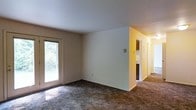 This single-floor residence offers a straightforward and functional layout designed for comfortable everyday living. The main living area is finished with wall-to-wall carpet and features clean, neutral-toned walls that create a bright and open atmosphere. A sliding glass door allows natural light to pour into the space while offering a pleasant view of the surrounding greenery outdoors, adding a connection to nature from within the home. The open wall space provides ample flexibility for furniture arrangement and personalization. Adjacent to the living area, a partial view into the kitchen reveals dark wood cabinetry, hinting at a practical and classic kitchen design. A hallway extends from this transitional area, suggesting access to additional rooms such as bedrooms and bathrooms beyond. The overall layout flows efficiently, with each zone connecting naturally to the next. The neutral color palette and simple finishes throughout give the space a clean, move-in-ready feel, providing a solid foundation for a variety of interior styles. The presence of natural light and greenery views contribute to a calm and livable ambiance throughout this single-level home.