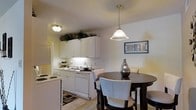 This single-floor residence offers a thoughtfully designed layout that blends functionality with a warm, welcoming ambiance. The kitchen is outfitted with crisp white cabinetry that extends to the upper walls, providing generous storage space. Upper cabinets reach toward the ceiling, maximizing vertical storage and lending a structured, organized feel to the space. The kitchen includes a white electric range, a dishwasher, and a sink, all neatly arranged along a clean, light-toned countertop. Under-cabinet lighting adds a subtle glow that enhances both the practicality and atmosphere of the cooking area. Adjacent to the kitchen, the dining area is illuminated by a elegant pendant light that hangs above a round dark-wood table surrounded by upholstered chairs, creating an inviting setting for meals. The light-toned flooring and neutral wall colors throughout contribute to an airy, open feel. Decorative touches, including wall art and potted greenery atop the upper cabinets, add character and a lived-in warmth to the space. The overall design emphasizes clean lines, ample storage, and a cohesive neutral palette that makes the space feel both practical and comfortable.