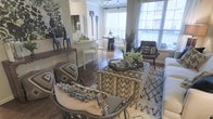 This single-floor residence showcases an open-concept design that connects the living, dining, and kitchen areas into one cohesive, flowing space. The living area anchors the layout with a textured, geometric-patterned ottoman serving as a central coffee table, surrounded by woven accent chairs adorned with fringe throw pillows, and a plush, intricately patterned area rug underfoot. Dark hardwood flooring runs throughout, providing a warm, grounding contrast to the light-toned walls.

The kitchen features a raised bar counter with white modern bar stools, offering a casual seating option and a natural visual divide between the kitchen and living spaces. Beyond the kitchen, the dining area is illuminated by a sleek track lighting fixture and benefits from generous natural light streaming through large windows. A glass-top dining table paired with contemporary chairs adds an airy, modern touch.

Large windows along the perimeter flood the space with daylight and offer views of an outdoor balcony area. Decorative elements, including sculptural figurines, botanical arrangements, and wall art, contribute to a layered, curated aesthetic throughout the home.