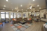 This single-floor community clubroom offers a thoughtfully designed open-concept layout that seamlessly blends lounge and dining areas into one cohesive gathering space. Wood-look plank flooring runs throughout, providing a warm, consistent foundation, while crown molding with dark accent trim adds a refined architectural detail to the neutral-toned walls and ceilings.

The central lounge area is anchored by a generously sized L-shaped sectional sofa in rich brown upholstery, accented with colorful throw pillows and centered over a bold, patterned area rug in teal, orange, and earth tones. A wall-mounted flat-screen television and a wooden bench occupy one wall, while an ornate sunburst mirror and dark console cabinet serve as a decorative focal point near the entry area. Multiple ceiling fans with integrated lighting are distributed throughout, ensuring comfort across the space.

The dining area is positioned near floor-to-ceiling windows and a glass sliding door, flooding the space with natural light and offering direct views of an outdoor pool and surrounding grounds. Round dining tables with upholstered chairs provide ample seating. An adjacent kitchenette is visible beyond the main area, completing the functional amenity space.