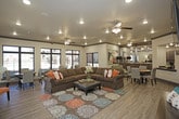 This single-floor community clubroom offers a thoughtfully designed open-concept layout that seamlessly blends lounge and dining areas into one cohesive gathering space. Wood-look plank flooring runs throughout, providing a warm, consistent foundation, while crown molding with dark accent trim adds a refined architectural detail to the neutral-toned walls and ceilings.

The central lounge area is anchored by a generously sized L-shaped sectional sofa in rich brown upholstery, accented with colorful throw pillows and centered over a bold, patterned area rug in teal, orange, and earth tones. A wall-mounted flat-screen television and a wooden bench occupy one wall, while an ornate sunburst mirror and dark console cabinet serve as a decorative focal point near the entry area. Multiple ceiling fans with integrated lighting are distributed throughout, ensuring comfort across the space.

The dining area is positioned near floor-to-ceiling windows and a glass sliding door, flooding the space with natural light and offering direct views of an outdoor pool and surrounding grounds. Round dining tables with upholstered chairs provide ample seating. An adjacent kitchenette is visible beyond the main area, completing the functional amenity space.