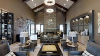 Located at 740 Bridgeport Ave in Shelton, Connecticut, this single-floor space makes a striking first impression with its grand, open-concept lobby. The soaring vaulted ceiling is accentuated by rich exposed dark wood beams and a stunning tiered crystal chandelier that serves as a central focal point, casting warm, inviting light throughout the space. A floor-to-ceiling stacked stone fireplace anchors the room, providing both a dramatic architectural statement and a cozy gathering atmosphere. The interior design blends contemporary and rustic elements seamlessly, with a sophisticated palette of charcoal grays, warm taupes, and gold accents reflected in the carefully curated furniture arrangement. Plush seating areas are thoughtfully positioned to encourage conversation and relaxation, complemented by decorative lamps, sculptural centerpieces, and tasteful artwork on the walls. Large glass-paneled doors and surrounding windows invite natural light deep into the interior, enhancing the open and airy feel of the space. Recessed lighting further highlights the architectural details, while warm wood-toned flooring grounds the design with a natural, welcoming quality. Overall, the space exudes a polished, upscale ambiance that is both functional and visually impressive.