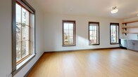 This well-designed single-floor unit offers a clean, modern aesthetic with light hardwood flooring and neutral tones carried throughout every room. The kitchen is a functional standout, featuring gray shaker-style cabinetry, white subway tile backsplash, stainless steel appliances including a refrigerator, electric range, microwave, and dishwasher, as well as open wooden shelving that adds a warm, contemporary touch. A window beside the kitchen brings in natural light, enhancing the space's bright and airy feel. The main living area is generously sized, with multiple windows framing views of the surrounding neighborhood, filling the room with abundant natural light. The clean white walls and open layout provide a versatile canvas for a variety of living arrangements. The bathroom is thoughtfully appointed with a wide vanity featuring a white countertop, undermount sink, and gray cabinetry, complemented by a stacked washer and dryer unit — a practical convenience integrated directly into the space. The entry foyer, visible from the kitchen corridor, features a classic wood-paneled door with a modern black hardware finish, offering a welcoming transition into the unit. Overall, the space balances modern finishes with functional design, making it both practical and inviting.