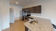 This well-appointed single-floor unit offers a thoughtfully designed layout that balances functionality and modern style throughout. The open-concept kitchen is a standout feature, equipped with stainless steel appliances including a refrigerator, electric range, and over-the-range microwave. Rich, dark-toned cabinetry pairs elegantly with granite countertops, while a subway tile backsplash and a peninsula sink add both practicality and visual appeal. Light wood-style flooring flows seamlessly through the kitchen and into the living area, creating a cohesive and inviting atmosphere. The living space is bright and airy, benefiting from a large sliding glass door that opens onto a private balcony, offering outdoor views and ample natural light. Vertical blinds provide flexible light control throughout the day. The bedroom is generously sized, featuring plush carpet flooring, multiple windows for natural light, and sufficient space for furnishings. A dedicated storage dresser is included, enhancing the room's practicality. The bathroom is clean and contemporary, outfitted with a granite-topped dark vanity, a toilet, and brushed nickel hardware accents including a towel bar and ring. The unit's neutral white walls and consistent finishes throughout contribute to a fresh, modern ambiance.