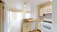 This single-floor space showcases a practical, open-concept layout that seamlessly connects the kitchen and living area. The kitchen is equipped with white cabinetry offering generous storage, a light-toned countertop, a double-basin sink, a white gas range, and a dishwasher. A peninsula-style counter serves as a natural divider between the kitchen and the adjoining living space, enhancing the open flow while maintaining functional separation. Recessed cylindrical ceiling-mounted light fixtures illuminate the kitchen area. The living space beyond features neutral carpet flooring and benefits from a large window dressed with vertical blinds, allowing natural light to filter in and brighten the interior. The walls and ceilings are finished in crisp white tones throughout, contributing to a clean and airy atmosphere. The textured ceiling adds a subtle design detail consistent with the space's understated aesthetic. Overall, the layout prioritizes functionality and openness, making efficient use of the available square footage.