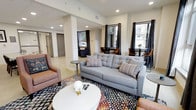 This single-floor space presents a well-appointed communal lounge area defined by a clean, contemporary design. The open-plan layout feels generously proportioned, with structural columns subtly dividing the space into distinct functional zones without compromising the overall sense of openness. Recessed lighting throughout the ceiling provides consistent, even illumination that complements the abundant natural light streaming in through multiple windows dressed with dark curtain panels, offering an attractive contrast to the bright, neutral walls and light-toned flooring. The main seating area is anchored by a tufted grey sofa adorned with striped accent pillows, paired with warm-toned upholstered armchairs and a round white coffee table atop a bold, multicolored patterned area rug that adds visual energy to the room. Toward the perimeter, bistro-style tables with wooden chairs are positioned near the windows, creating cozy nooks suited for quiet conversation or individual use. In the background, additional spaces — including what appears to be a private room or alcove — extend the functionality of the floor. The overall ambiance is modern yet welcoming, combining practical furnishings with thoughtful design elements that make the space both versatile and comfortable.
