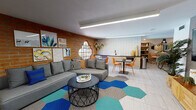 This single-floor community space blends industrial character with modern, colorful design to create a welcoming and versatile gathering area. The main lounge is anchored by a striking full-length exposed brick wall, against which a large sectional sofa is arranged atop a bold hexagonal-patterned area rug in shades of blue, teal, and gray. A curated gallery wall of eclectic artwork, a round porthole-style window, and linear LED ceiling fixtures add visual interest and warmth throughout the space.

Adjacent to the lounge, a well-appointed kitchenette features light wood cabinetry with dark countertops, a matte black sink, an under-counter refrigerator, a microwave, and a bold geometric-patterned tile backsplash. Diamond-shaped wall planters and woven decorative accents add a natural touch. A compact dining table with vibrant orange chairs provides a casual spot for meals or conversation.

A dedicated game room offers a foosball table as its centerpiece, flanked by built-in wood shelving stocked with board games and décor. A teal accent wall, wire pendant lights, and a cozy built-in bench seating nook complete the playful yet refined atmosphere.

The restroom continues the design language with a painted white brick wall, a globe pendant light, dark wood-slat platform flooring, and a modern vanity with matte black fixtures and a concrete-style countertop.