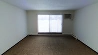 This single-floor space offers a straightforward and functional layout designed for comfortable everyday living. The main living area features wall-to-wall carpeting in a neutral tan tone, providing a warm and understated foundation throughout the room. A large sliding glass door serves as the focal point of the space, flanked by vertical blinds that allow for adjustable light control and privacy. The door provides an abundance of natural light, brightening the interior and offering a view to the outdoors. Climate comfort is well addressed with a wall-mounted air conditioning unit positioned above the sliding door, complemented by baseboard heating that runs along the perimeter of the room, ensuring year-round temperature regulation. The walls are finished in a clean, neutral off-white tone, and the textured ceiling adds a subtle architectural detail. The open, uncluttered layout of the living area provides flexibility for a variety of furniture arrangements, making it easy to personalize the space. Overall, the room presents a clean, move-in ready environment with practical features suited for daily living.