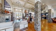 Located at 401 Old Pleasant Grove Rd in Mount Juliet, Tennessee, this single-floor community space offers a generously sized and thoughtfully designed interior. The grand entryway welcomes visitors with soaring double-height ceilings, floor-to-ceiling glass panel doors, and stacked stone columns that frame the space with rustic elegance. Polished stained concrete floors run throughout, providing a warm, earthy tone that complements the overall design aesthetic.

The expansive living area serves as the heart of the space, anchored by a striking floor-to-ceiling stacked stone fireplace topped with a large-screen television. Coffered ceilings adorned with elegant pendant chandeliers add architectural sophistication, while large transom windows flood the room with natural light and offer views of the outdoors. Comfortable, stylishly arranged seating areas invite gathering and relaxation.

The kitchen is designed for functionality and style, featuring dark espresso cabinetry, granite countertops, a mosaic tile backsplash, and a full suite of stainless steel appliances. The galley-style layout provides ample counter space and flows seamlessly into the adjoining living and dining areas. Additional seating nooks and a billiards area round out this versatile, well-appointed community space.