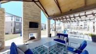 This covered outdoor lounge offers a well-appointed gathering space designed for both relaxation and socializing. The structure is anchored by a striking floor-to-ceiling stacked stone fireplace, which serves as the focal point of the space and includes a mounted flat-screen television above the mantle, making it ideal for entertaining. The vaulted ceiling features warm wood paneling supported by exposed beams, creating an inviting, lodge-inspired atmosphere while providing ample shelter. The open-air design allows natural light and outdoor breezes to flow freely through the space, maintaining a seamless connection to the surrounding exterior environment. Comfortable seating with bold blue cushions and coordinating accent pillows is arranged around a central coffee table atop a patterned area rug, offering a cohesive and inviting setup. A pergola-covered extension adjacent to the main structure provides additional outdoor seating and appears to include grilling amenities, further enhancing the space's functionality. Views of the surrounding community and pool area are visible from within the lounge, reinforcing its social and communal character. The overall ambiance is modern yet warm, balancing stylish design with practical outdoor living.
