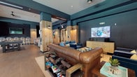 This single-floor amenity space in Atlanta, GA, 30318, showcases a cohesive industrial-modern design defined by deep teal and charcoal walls, reclaimed wood-clad columns, and large-format tile flooring throughout. The layout flows seamlessly between several distinct zones, creating a versatile environment for both relaxation and socializing.

The media lounge anchors one section of the space, furnished with a substantial tufted leather sofa, navy blue accent chairs, and a wall-mounted flat-screen display. A rustic coffee table stocked with board games adds a casual, communal touch. Adjacent to this area, wood-paneled double glass doors lead to additional rooms, with colorful artwork punctuating the warm natural wood surfaces along the corridor.

A dedicated game room features a full-size billiards table, casual dining tables with mixed seating, and vibrant abstract artwork on the teal walls. Pendant lighting with black metal shades provides focused illumination, while a ceiling fan adds comfort overhead.

Floor-to-ceiling glass walls and large windows flood portions of the space with natural light and frame views of mature trees and an outdoor pool area. High-top bistro tables positioned near the glass provide a bright transition point between indoors and the exterior. Throughout the space, reclaimed wood accents, industrial sconce lighting, and carefully curated décor create a cohesive, inviting atmosphere.