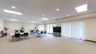 This community room is a well-appointed, single-floor multipurpose space designed to accommodate a variety of events and gatherings. The room features a drop ceiling with recessed lighting and skylights that bring in natural light, creating a bright and welcoming atmosphere. Large windows and a sliding glass door along one wall further enhance the sense of openness while offering views of the surrounding outdoor area. The flooring is covered in neutral-toned carpet, contributing to a clean and functional aesthetic throughout. The space is furnished with multiple rectangular tables and black chairs arranged in a flexible layout, making it easy to reconfigure for different uses, from meetings and workshops to social events. A large flat-panel display screen mounted on a wheeled stand serves as a central presentation focal point, supporting a range of multimedia needs. One side of the room features a room divider with vertical blinds, suggesting the space can be sectioned off as needed. The walls carry a subtle textured finish in light tones, and decorative plants add a touch of greenery. Overall, the community room offers a practical, adaptable environment well-suited for group activities and community programming.