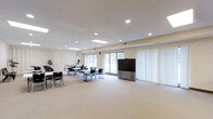 This community room is a well-appointed, single-floor multipurpose space designed to accommodate a variety of events and gatherings. The room features a drop ceiling with recessed lighting and skylights that bring in natural light, creating a bright and welcoming atmosphere. Large windows and a sliding glass door along one wall further enhance the sense of openness while offering views of the surrounding outdoor area. The flooring is covered in neutral-toned carpet, contributing to a clean and functional aesthetic throughout. The space is furnished with multiple rectangular tables and black chairs arranged in a flexible layout, making it easy to reconfigure for different uses, from meetings and workshops to social events. A large flat-panel display screen mounted on a wheeled stand serves as a central presentation focal point, supporting a range of multimedia needs. One side of the room features a room divider with vertical blinds, suggesting the space can be sectioned off as needed. The walls carry a subtle textured finish in light tones, and decorative plants add a touch of greenery. Overall, the community room offers a practical, adaptable environment well-suited for group activities and community programming.