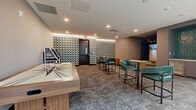 This well-appointed entertainment lounge offers a dynamic and social atmosphere designed for recreation and relaxation. The single-floor space is thoughtfully organized into distinct activity zones across a generously sized open layout, all unified by sophisticated patterned carpet flooring and recessed lighting throughout.

A standout feature is the TruGolf sports simulator, enclosed by dark draping and a large projection screen, allowing guests to enjoy golf, soccer, and other virtual sports on an artificial turf platform. Adjacent to the simulator, a dedicated games area includes a solid wood shuffleboard table, a classic Atari arcade cabinet, and framed vintage-style wall art that adds a nostalgic touch.

The lounge area is anchored by teal velvet barrel chairs and high-top tables with bar-height seating, creating inviting spaces for socializing. A wet bar with teal mosaic tile backsplash and an under-counter refrigerator provides convenient refreshment service. Floor-to-ceiling windows and a sliding glass door flood this area with natural light and open directly onto an outdoor courtyard, seamlessly blending indoor and outdoor living.

A bold geometric patterned feature wall frames the main entrance, adding a striking visual focal point. The overall design palette — combining warm neutrals, deep teals, and rich wood tones — strikes a balance between modern sophistication and casual entertainment appeal.