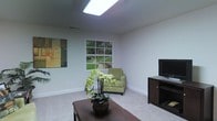 This single-floor space features a thoughtfully designed living area that combines natural light with a clean, neutral aesthetic. A prominent skylight installed in the ceiling serves as a key architectural feature, flooding the room with abundant overhead light and enhancing the sense of openness. Complementing the skylight, a multi-pane window provides views of a lush green outdoor setting, connecting the interior to the surrounding landscape and bringing in additional natural light.

The walls are finished in a soft, neutral gray-green tone that contributes to a calm and airy ambiance. Light-colored carpeting covers the floor, adding warmth underfoot and reinforcing the room's subdued color palette. The space accommodates a comfortable seating arrangement, including a green upholstered armchair positioned near the window, alongside a dark wood coffee table and a matching media console that houses a flat-screen television. A colorful abstract artwork adorns one wall, providing a visual focal point. Visible HVAC venting integrated into the ceiling and walls reflects practical, functional design throughout the space.