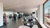 Located at 829 Garfield Ave in Jersey City, NJ, this modern fitness center offers a thoughtfully designed, single-floor exercise space that is both functional and visually dynamic. The open floor plan provides ample room for a diverse range of workout equipment, including a row of treadmills and elliptical machines positioned along floor-to-ceiling windows that flood the space with natural light and offer views of the surrounding outdoor scenery. The strength training area is well-appointed with cable machines, weight benches, a dumbbell rack, kettlebells, medicine balls, and a stability ball, catering to a full spectrum of fitness needs. A large wall of mirrors along one side of the room enhances the sense of space and allows users to monitor their form. Rubberized flooring throughout provides durability and safety underfoot. Recessed linear LED lighting runs along the ceiling, creating a bright and energizing atmosphere. A standout feature is a bold, colorful mural depicting the Statue of Liberty lifting a barbell, accompanied by the motivational phrase "Walk In Strong, Walk Out Stronger," adding a distinctly local and spirited character to the space. The gym also includes a water fountain near the entrance, ensuring convenience for all users.