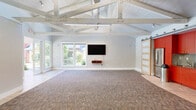 Located at 2925 Monument Blvd in Concord, CA, this single-level multipurpose space offers a generously open layout ideal for gatherings, meetings, or community events. The room's most striking architectural feature is its dramatic vaulted ceiling with exposed white-painted structural beams and rafters, lending the space a bright, airy character well above standard ceiling heights.

The interior is finished with neutral light-gray walls and a multi-tone commercial carpet that anchors the main floor area, while tile flooring transitions near the entryways and kitchen zone. A wall-mounted flat-screen television provides a functional focal point, accompanied by a small media shelf. Sliding barn-style doors reveal a fully equipped kitchen alcove outfitted with bold red cabinetry, stainless steel appliances including a refrigerator, gas cooktop, and oven, making the space well-suited for catered events.

Ample natural light floods the room through multiple sets of large windows and glass doors on opposing walls. One set of French doors opens directly to an outdoor pool area, offering seamless indoor-outdoor connectivity. Stacked folding chairs and a movable room divider panel system add flexible configuration options. Track and fluorescent lighting ensure well-distributed illumination throughout, completing this adaptable and inviting community venue.