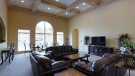 This distinguished single-floor residence showcases exceptional architectural craftsmanship throughout. The centerpiece of the home is a sweeping living room defined by soaring coffered ceilings with decorative molding and recessed lighting, lending a sense of grandeur to the space. A dramatic arched window floods the room with natural light, offering views of the surrounding neighborhood while creating a bright and airy ambiance. Warm, neutral-toned walls complement the classic design, and a graceful arched alcove adds an elegant architectural accent. The living room is generously proportioned, comfortably accommodating a full suite of dark leather sofas arranged around a wooden coffee table, with a media console and flat-screen television completing the entertainment area. The open layout promotes a natural flow through the space, while the interplay of natural and recessed lighting ensures the interior remains inviting at all hours. The overall design aesthetic blends traditional elegance with comfortable livability, making this home a refined and functional retreat suited for both everyday living and entertaining.