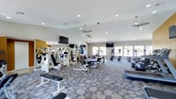 This fitness center is a spacious, single-floor facility designed to support a comprehensive workout experience. The room is finished with a stylish patterned carpet in neutral tones of grey and blue, providing both comfort and durability underfoot. The layout is thoughtfully organized, with strength training equipment — including cable machines, weight benches, and multi-function resistance stations — positioned on one side, while cardio equipment such as ellipticals, upright bikes, and stair climbers line the opposite side near the windows.

The ceiling is white and features recessed lighting alongside multiple ceiling fans, ensuring the space remains well-lit and well-ventilated throughout. Two flat-screen televisions are mounted at strategic points around the room, allowing members to stay entertained during their workouts. An accent wall painted in a warm golden-yellow hue adds a pop of color and visual interest to the space.

Large windows and glass doors along the far wall flood the interior with natural light and offer views to the outdoors, enhancing the open and airy ambiance of the facility. The overall design strikes a balance between functionality and comfort, making this fitness center a welcoming environment for users of all fitness levels.
